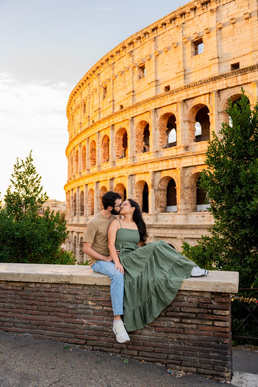 Couple kissing on a stone wall with the golden-lit Colosseum behind them at sunset in Rome