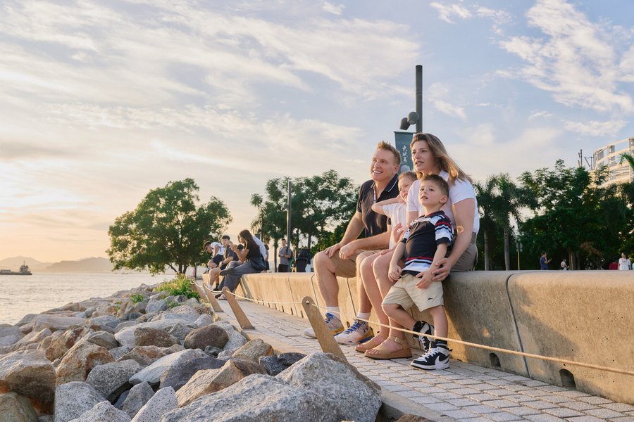 Family sitting on waterfront wall at golden hour with rocky shore and harbor view in Hong Kong