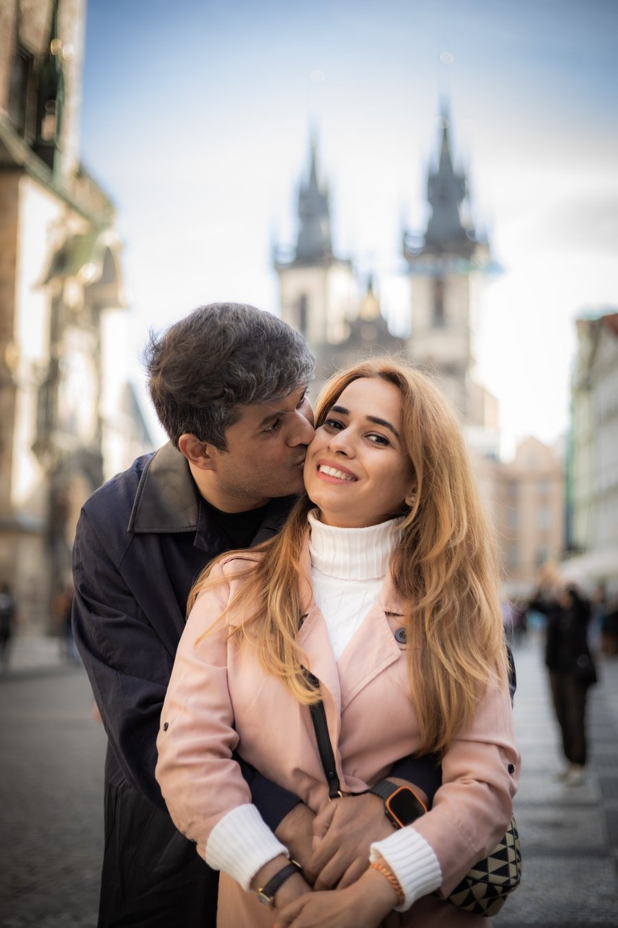 Couple embracing in Prague's Old Town Square with twin church spires in soft daylight