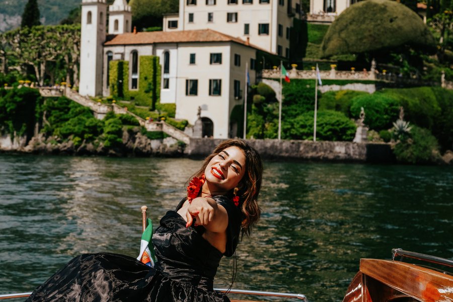 Woman with red earrings on a boat at Lake Como, Italian villa and gardens in bright daylight behind