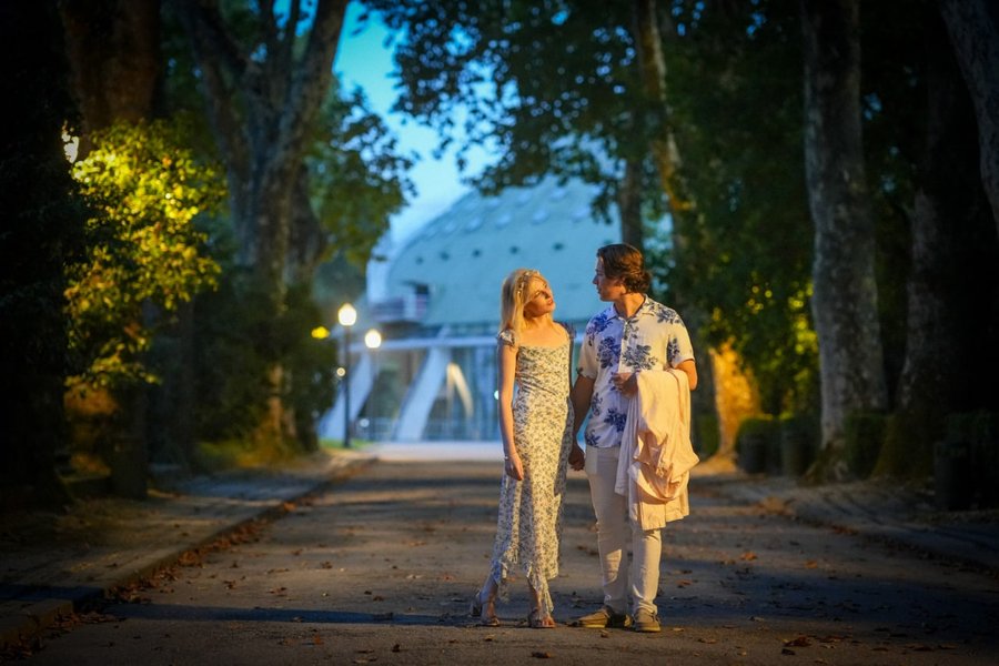 couple walking at dusk through tree-lined avenue with historic building beyond, golden hour light