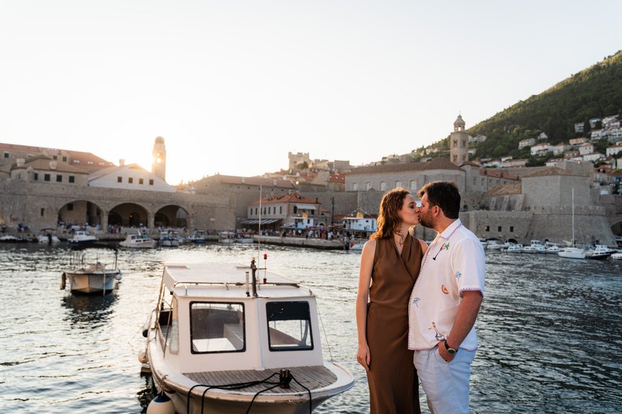 Couple kissing by a boat at sunset with Dubrovnik's historic walled city and harbor in the background