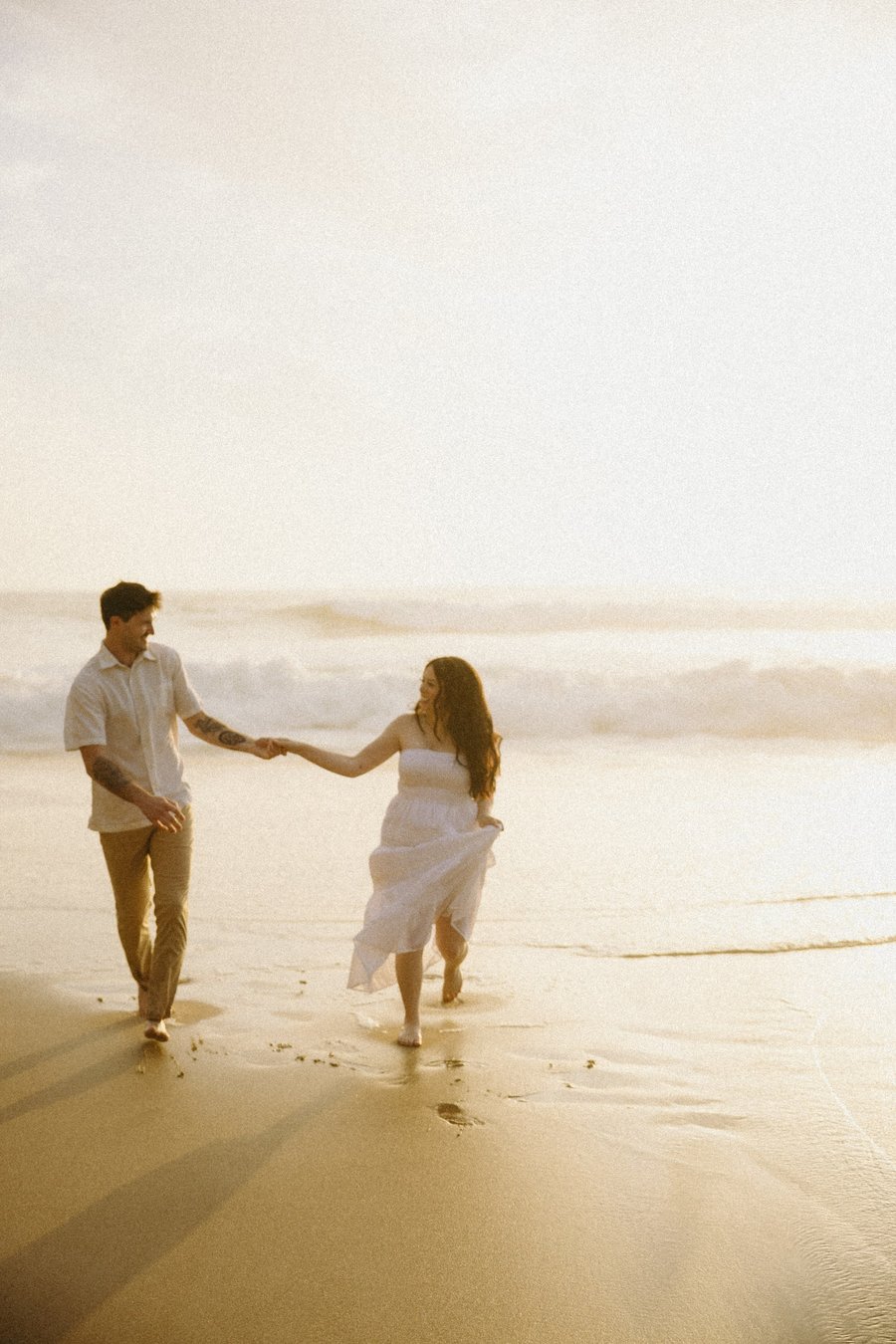 Couple holding hands on a sandy beach at golden hour, waves in the background