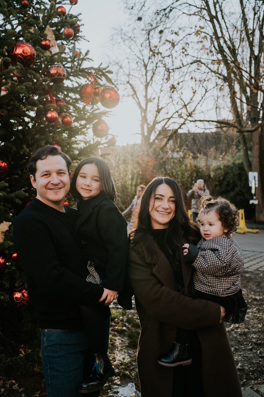 Family of four smiling under red ornaments on decorated evergreen tree at golden hour