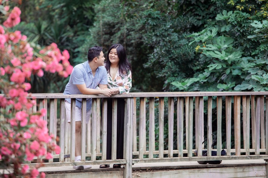 Couple standing on wooden bridge surrounded by lush green foliage and pink flowers in daylight
