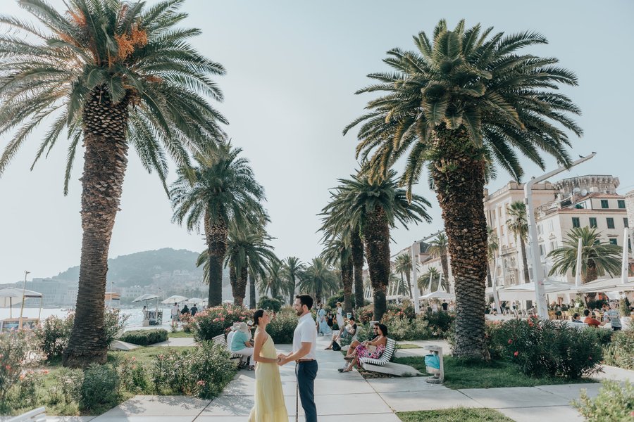 Couple standing among palm trees on a waterfront promenade in Split, with historic buildings and Mediterranean sea beyond