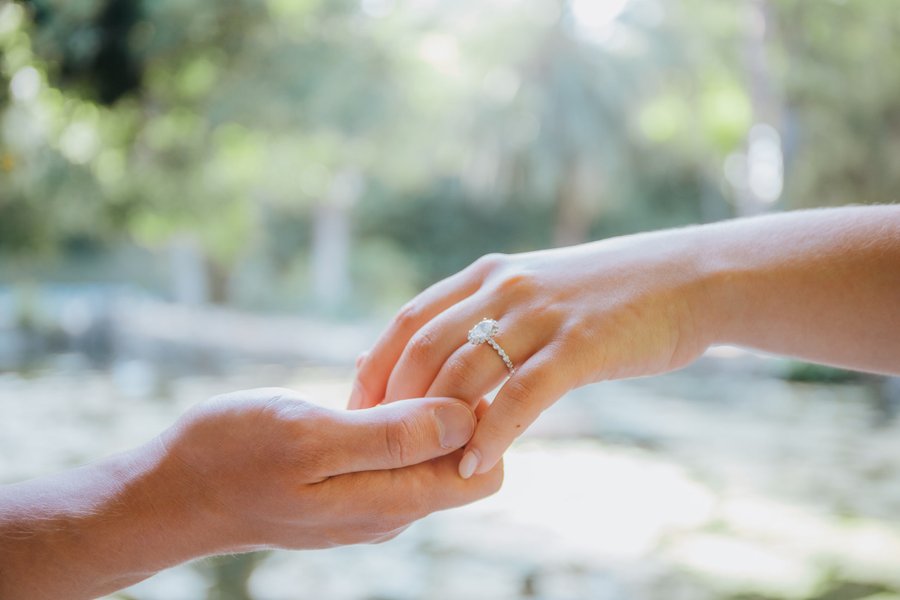 Couple holding hands with diamond engagement ring, lush green garden backdrop in daylight