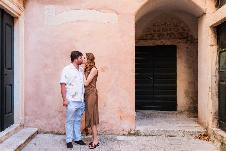 Couple posing against weathered pink stone walls in a historic Mediterranean alleyway during daytime