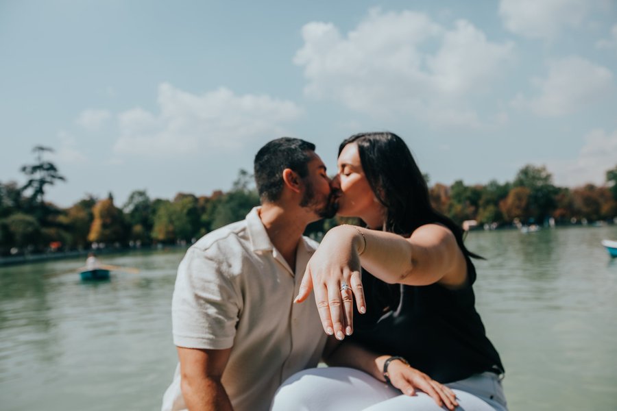 Couple kissing by a calm river with tree-lined banks and blue sky with white clouds