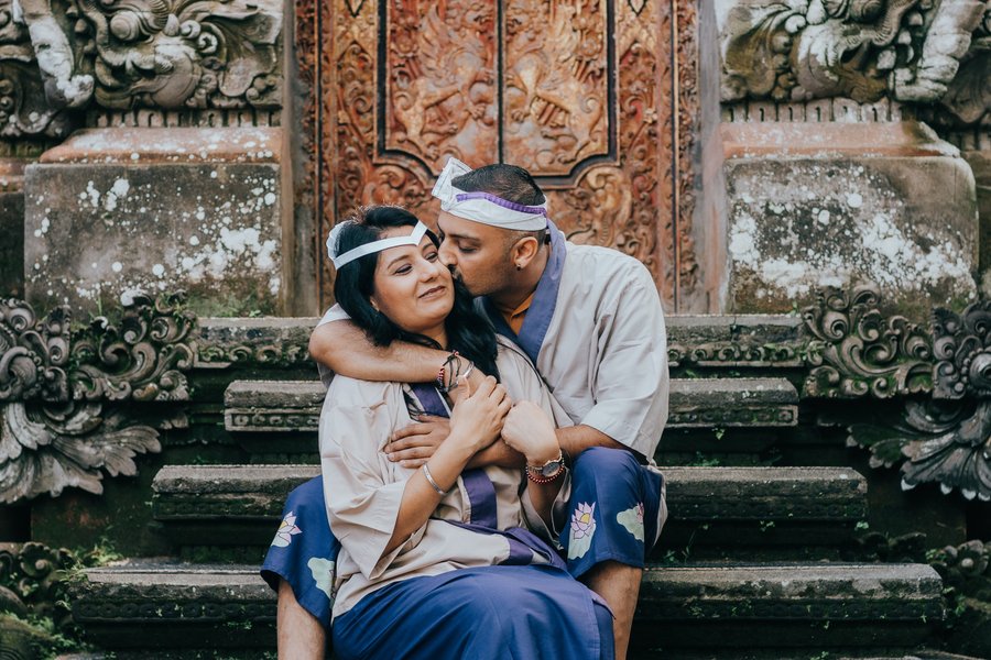 Couple in colorful headbands embrace on weathered stone steps before ornate carved wooden temple doors in Ubud