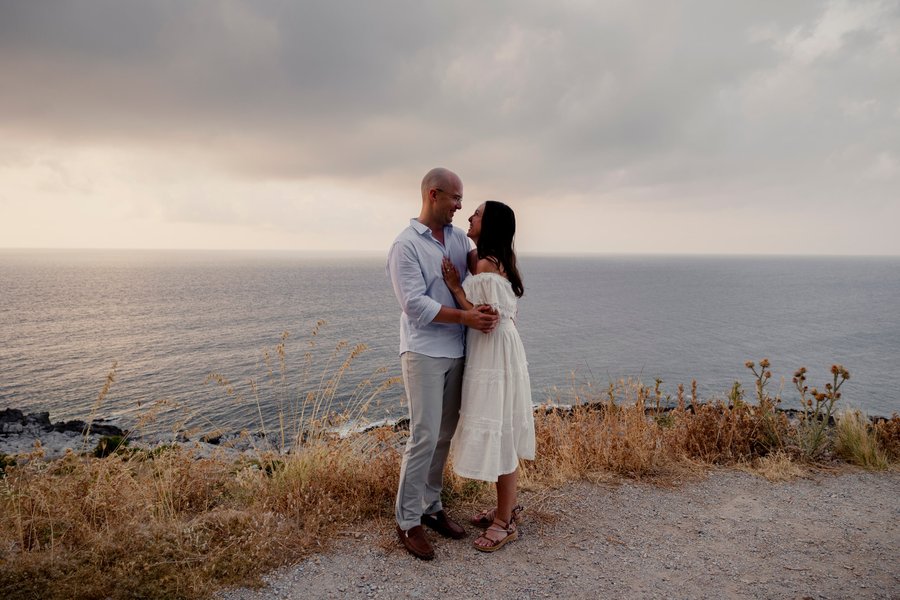 Couple embracing on a clifftop overlooking the sea at golden hour, dry coastal vegetation surrounding them