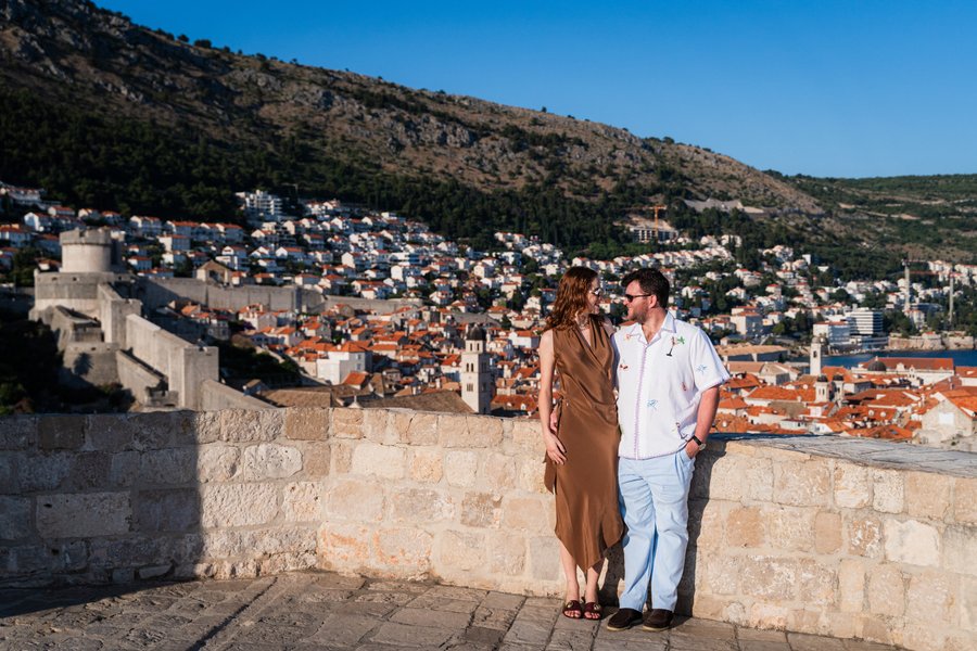 Couple standing on Dubrovnik's ancient city walls overlooking terracotta-roofed Old Town and Adriatic coastline under clear blue sky
