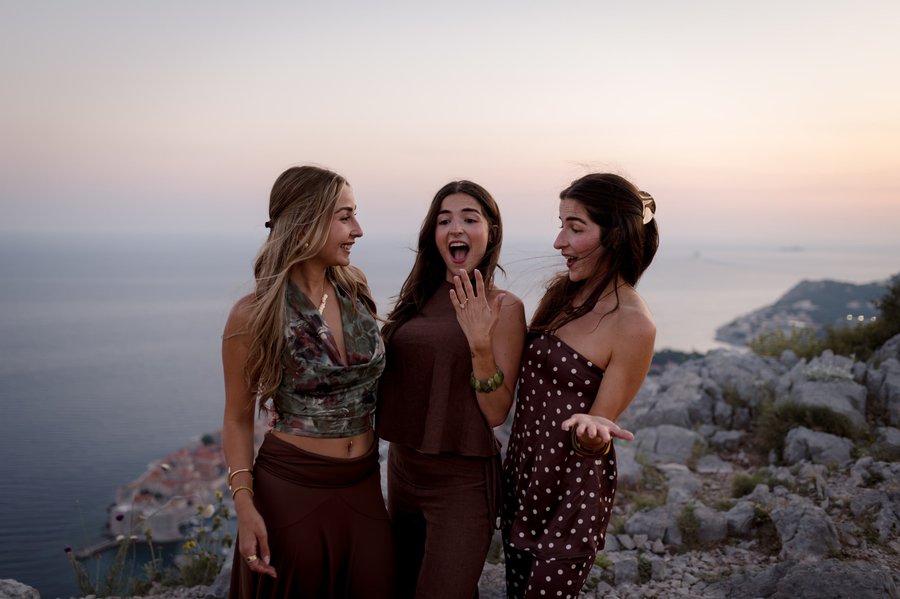 Three women laughing together at a rocky coastal viewpoint during sunset in Dubrovnik
