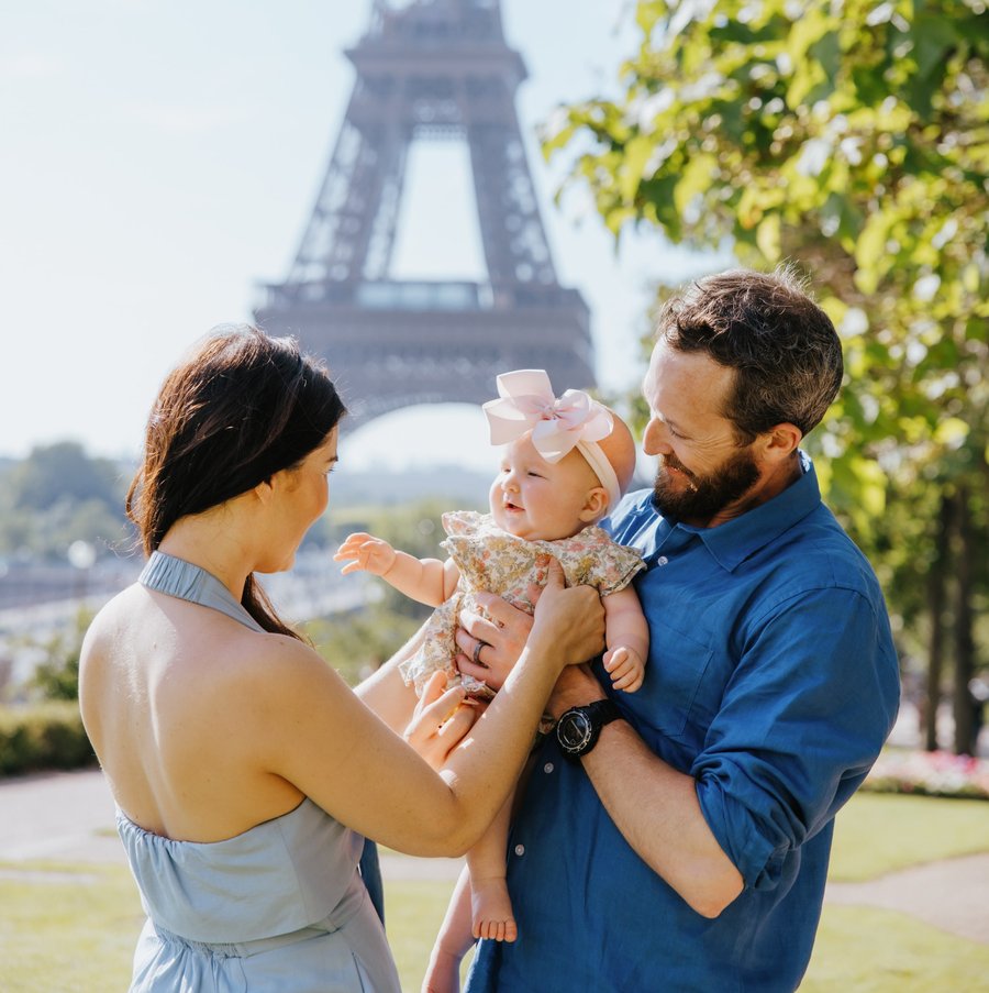 Family with infant in pink bow near Eiffel Tower on sunny day with green foliage
