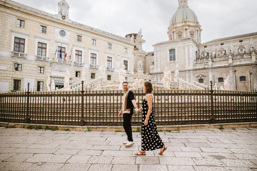 Couple walking past historic Sicilian buildings and fountain at Fontana Pretoria, Palermo on overcast day