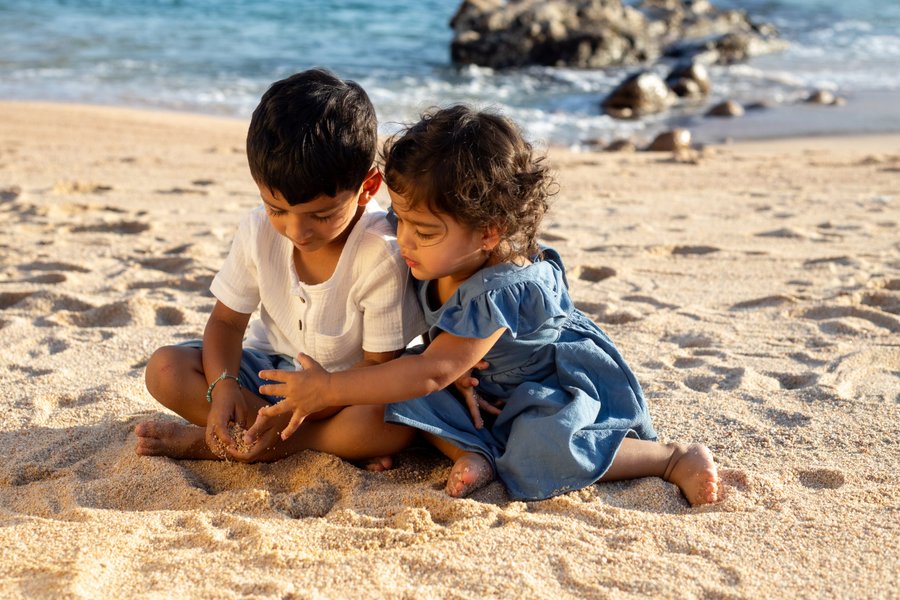 Two young children playing in the sand on a sunny beach with rocky outcrops and turquoise water behind them