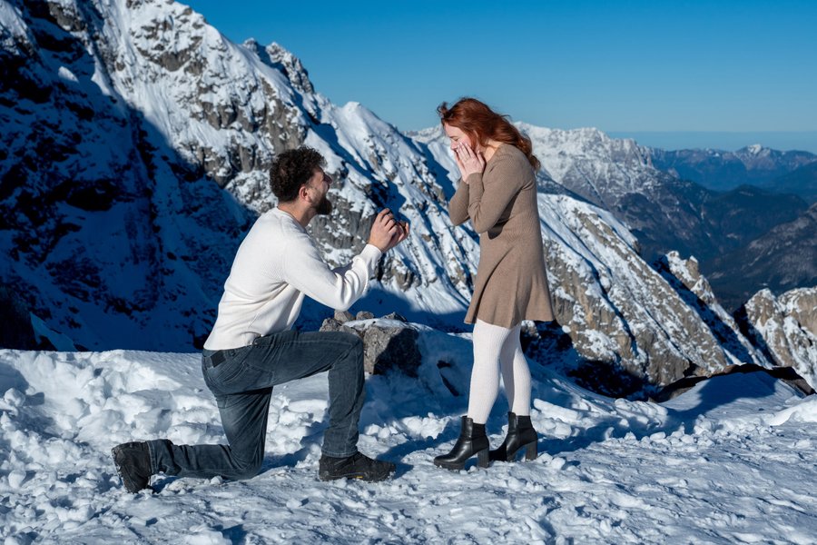 Man kneeling in snow proposing to smiling woman on snowy Innsbruck mountain ridge