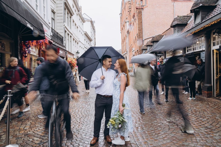 Couple with umbrella on rainy cobblestone street in historic Bruges, surrounded by pedestrians and traditional brick architecture