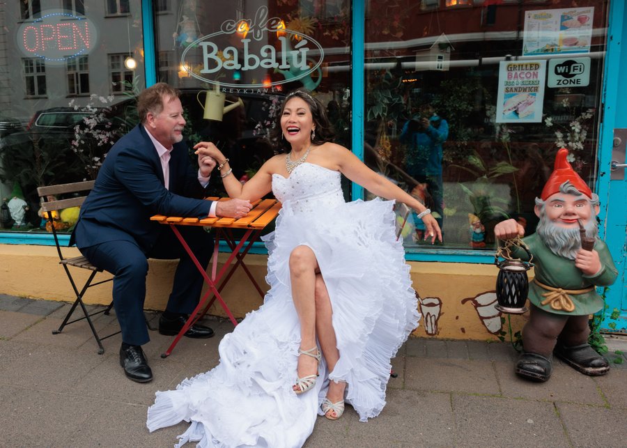 Bride in white wedding dress laughing with groom in blue suit outside colorful Babalu restaurant storefront in Reykjavík
