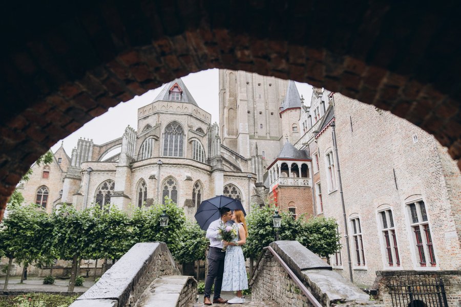 Couple with black umbrella on stone bridge framed by archway, Gothic cathedral behind, overcast day in Bruges