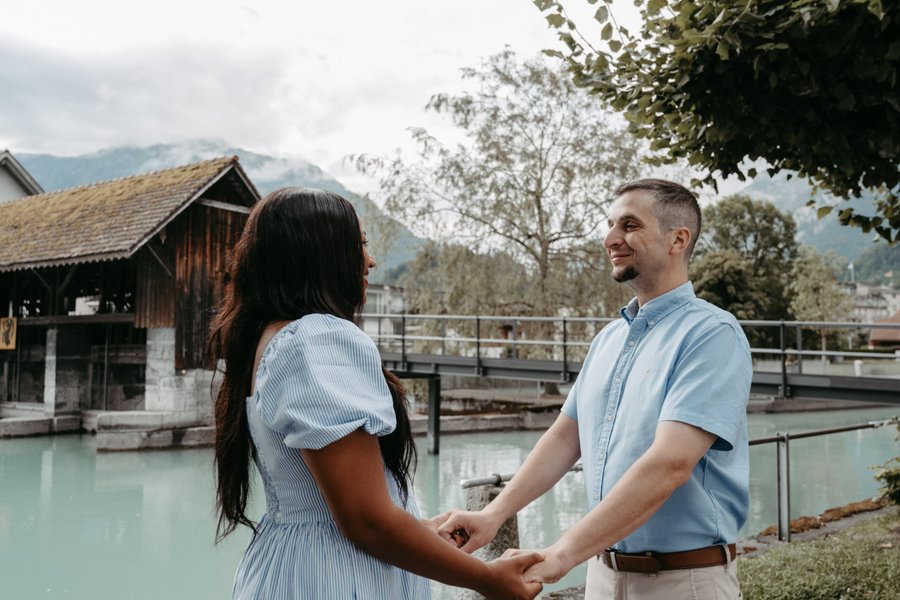 Couple holding hands by a turquoise river and wooden boathouse with mountains in the background