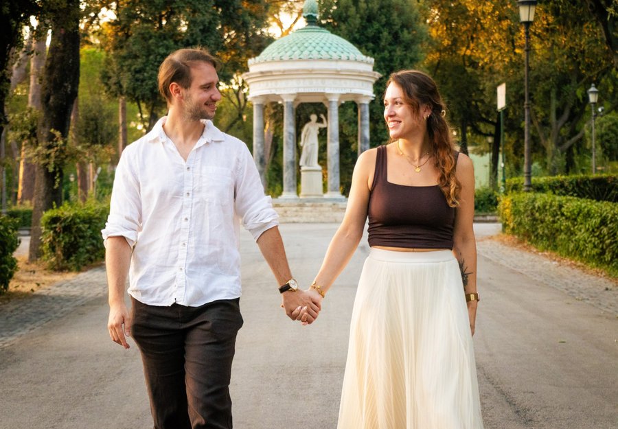 Couple holding hands walking through Villa Borghese gardens at golden hour with classical gazebo in background