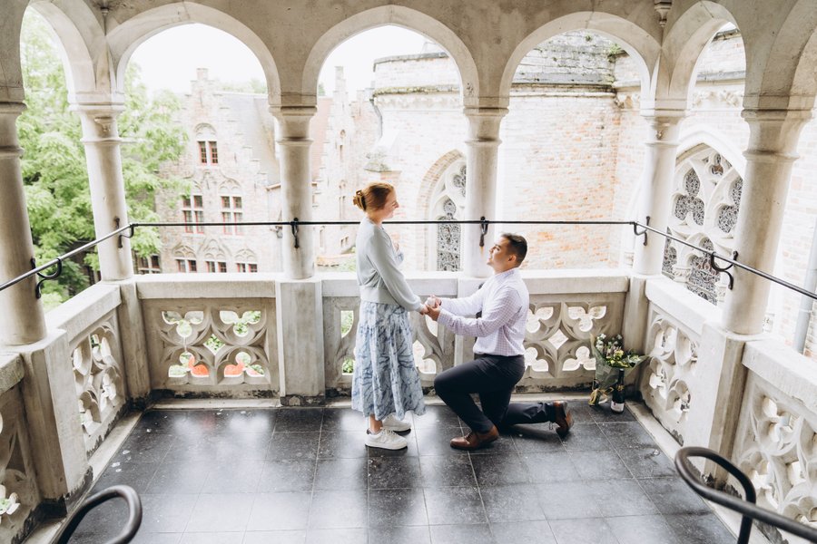 Man on one knee proposing to woman on stone balcony with Gothic arches and cathedral views in Bruges