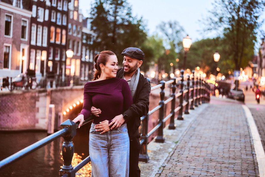 Couple smiling on a canal bridge at dusk, historic buildings and golden streetlights lining the waterway