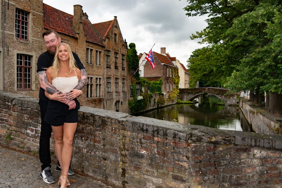 Couple posing on stone bridge over canal with medieval brick buildings and British flag in background, overcast day
