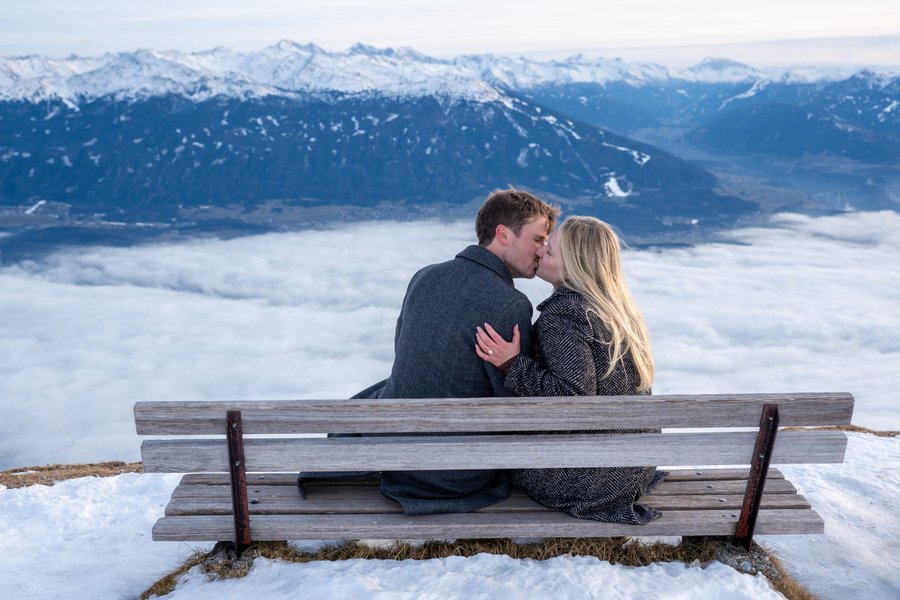 Couple kissing on a wooden bench above clouds, with snowy Alpine peaks behind them in Innsbruck