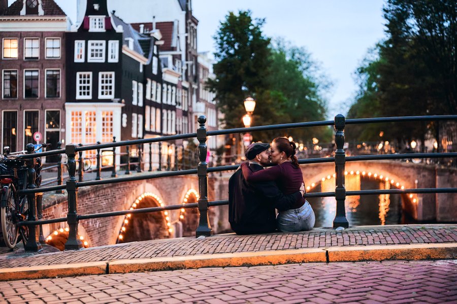 Couple sitting on a bridge over illuminated canal at dusk with historic Dutch architecture and bicycle nearby