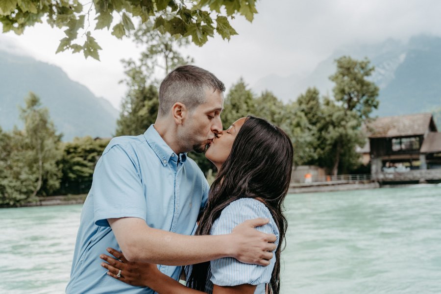 Couple kissing by turquoise river with alpine mountains and boathouse in background during daytime