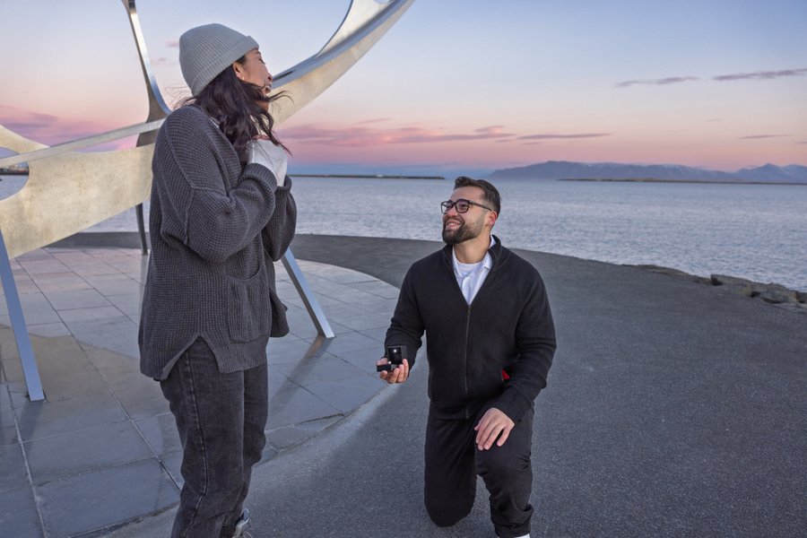 Couple at the Sun Voyager sculpture on a Reykjavík beach at sunset, with calm waters and mountains in the distance