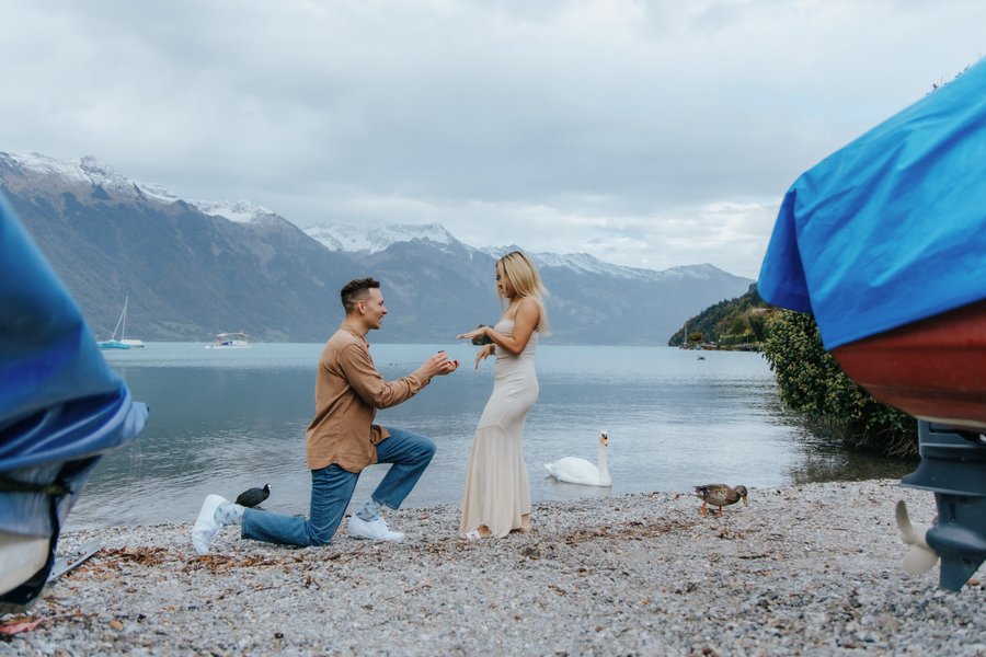 Man proposing to woman on Lake Brienz shore with snow-capped mountains and blue boats framing the scene