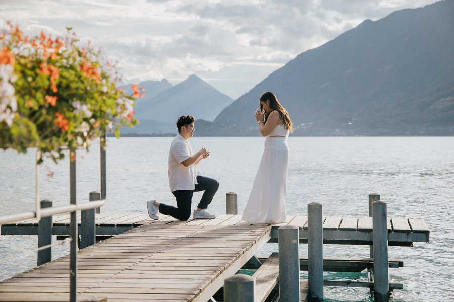 Man proposing to woman in white dress on wooden pier at Lake Brienz with mountains and orange flowers in background