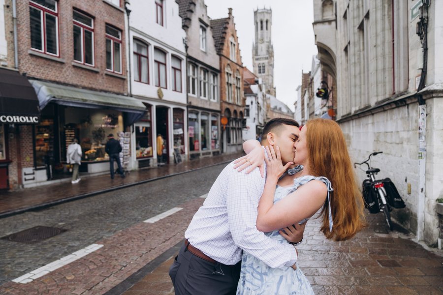 Couple kissing on wet cobblestone street lined with medieval architecture and belfry tower in bruges