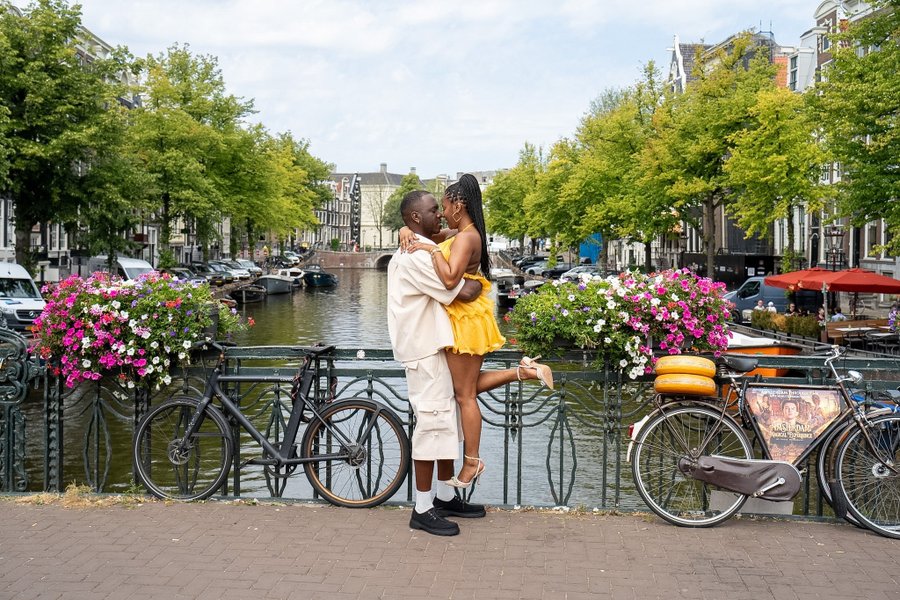Couple kissing by canal railing with bikes, pink flowers, and Amsterdam waterway lined with trees