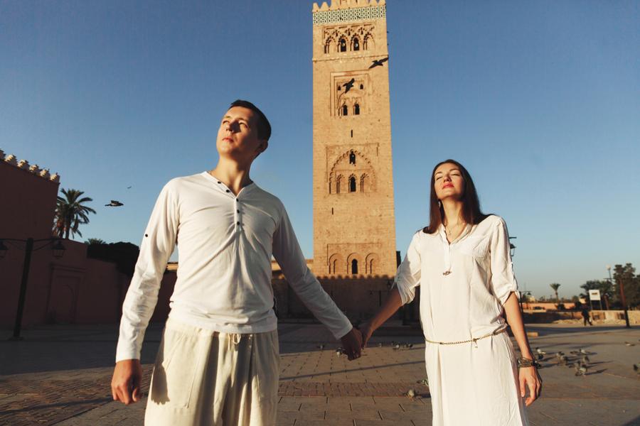 Couple holding hands in white outfits at the Koutubia Mosque in Marrakesh under clear blue sky